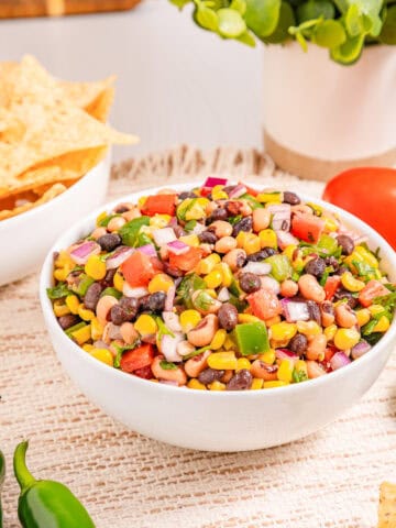 A bowl of colorful cowboy caviar salad with corn, tomatoes, and herbs, served next to tortilla chips and fresh vegetables.