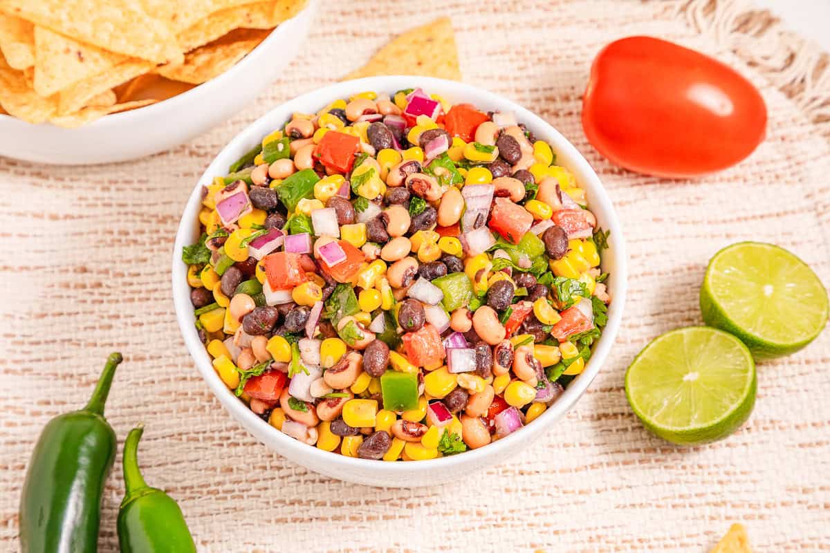 A bowl of colorful Cowboy Caviar, a zesty bean and corn salsa with chopped veggies, surrounded by chips, lime, tomato, and jalape&ntilde;os.