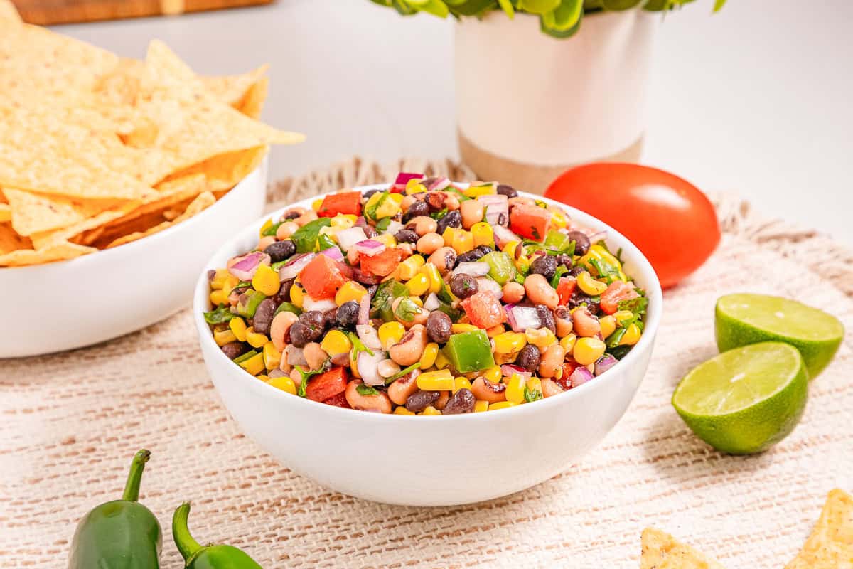 A bowl of colorful Cowboy Caviar with beans, corn, tomatoes, onions, and herbs sits beside tortilla chips, lime, and jalape&ntilde;o&mdash;a fresh take on the classic Cowboy Caviar Recipe.