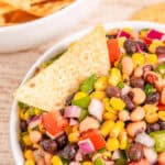 A bowl of colorful Cowboy Caviar salsa with a tortilla chip dipped in, and chips in a bowl in the background.