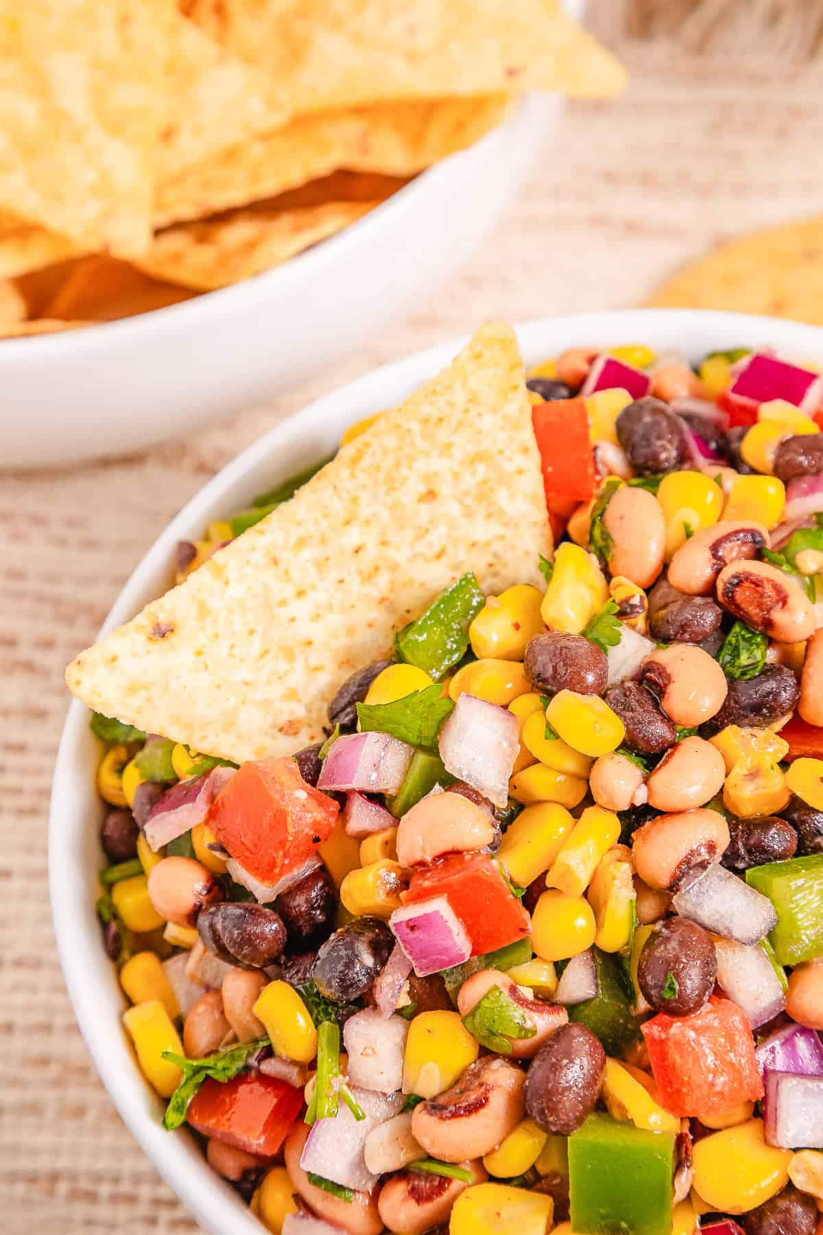 A bowl of colorful Cowboy Caviar salsa with a tortilla chip dipped in, and chips in a bowl in the background.