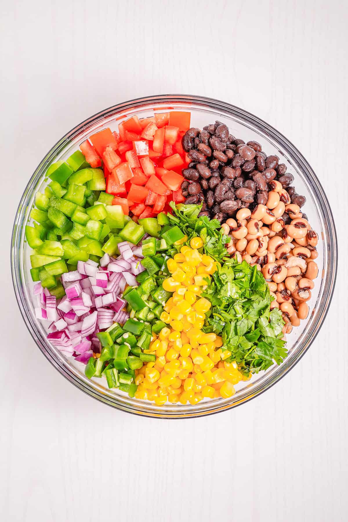 A glass bowl filled with chopped vegetables, beans, corn, and cilantro, arranged in colorful sections&mdash;perfect for serving a vibrant Cowboy Caviar recipe.