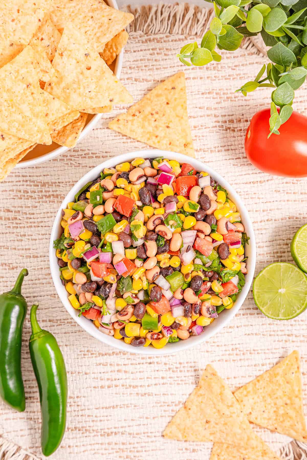 A bowl of colorful Cowboy Caviar with tortilla chips, jalape&ntilde;os, lime, and a tomato sits on a woven placemat&mdash;this vibrant dip recipe is perfect for snacking or parties.