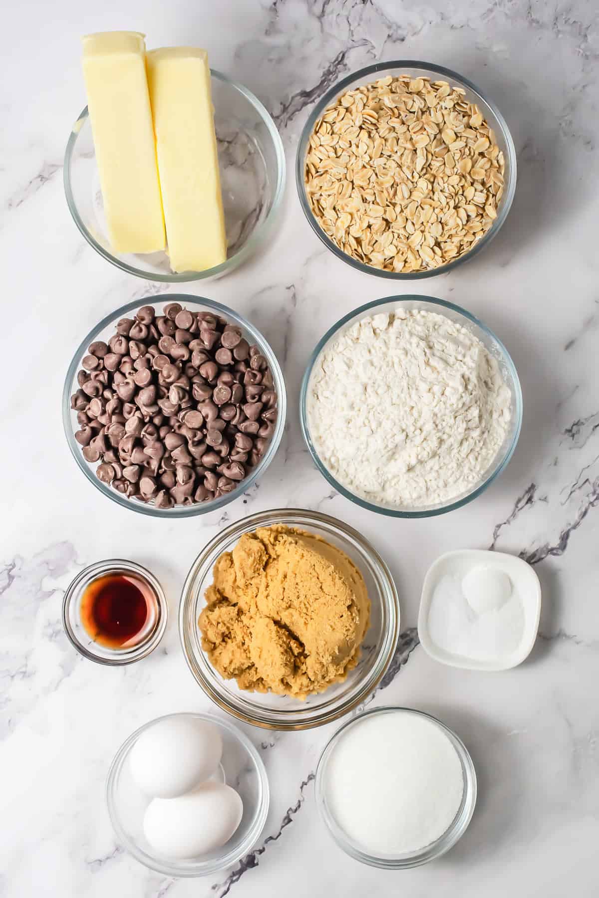 Ingredients for oatmeal bars on a marble surface: butter, oats, chocolate chips, flour, brown sugar, vanilla, eggs, sugar, and baking soda.