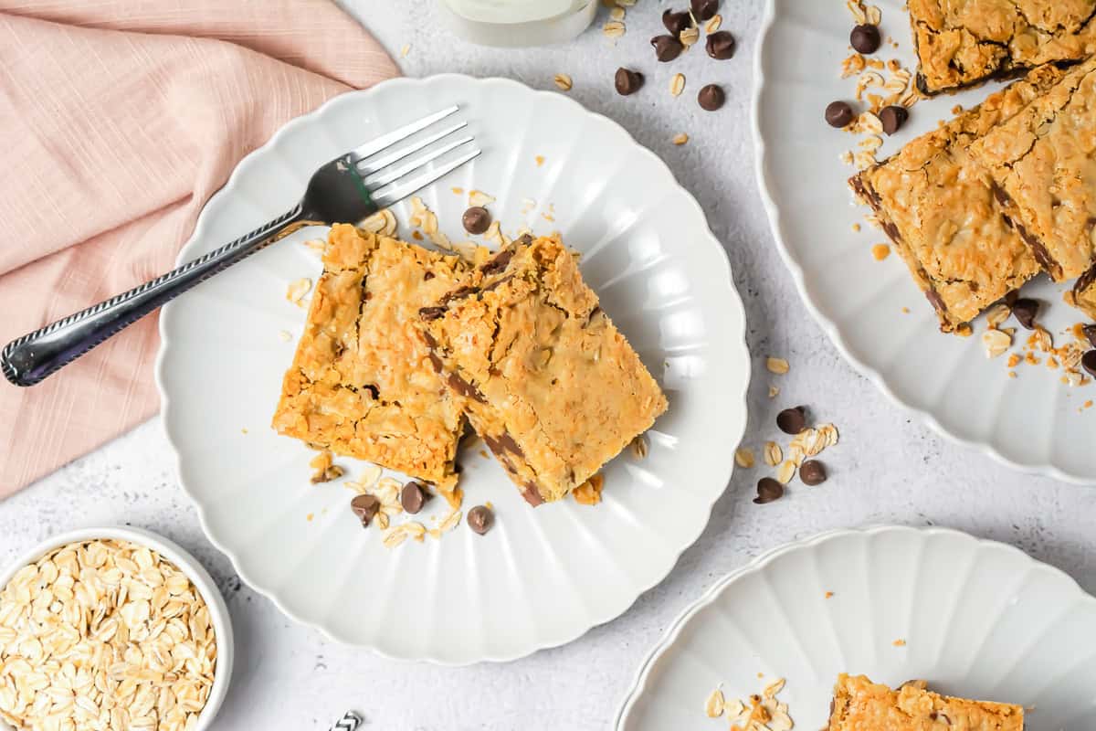 Two Oatmeal Chocolate Chip Bars rest on a white plate with a fork, surrounded by scattered oats and chocolate chips.