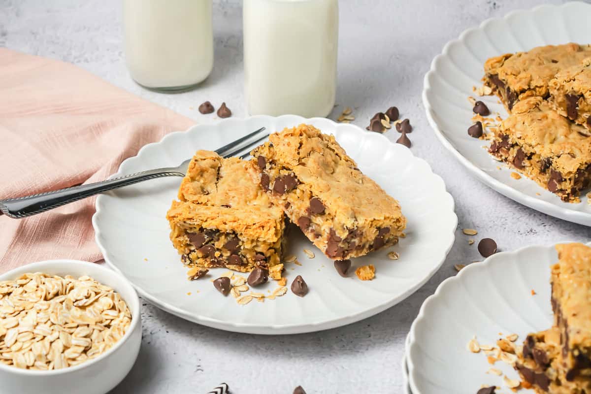 Two oatmeal chocolate chip bars rest on a white plate with forks, surrounded by oats and a glass of milk in the background.