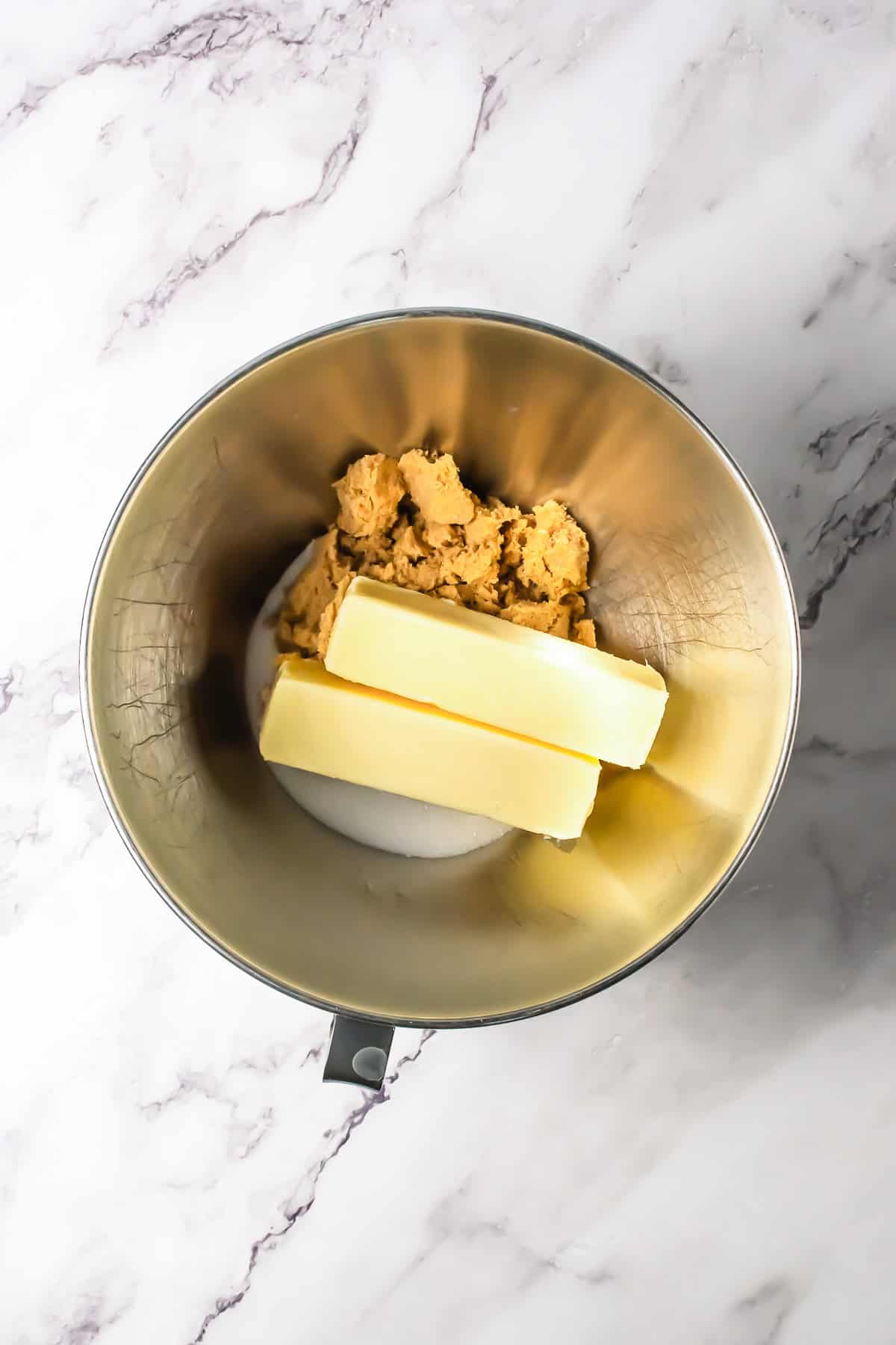 Two sticks of butter, brown sugar, and white sugar in a metal mixing bowl on a marble countertop&mdash;perfect for starting homemade oatmeal chocolate chip bars.