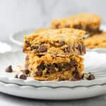 Two oatmeal bars packed with chocolate chips are stacked on a white plate, while more delicious bars and scattered chocolate chips tempt in the background.