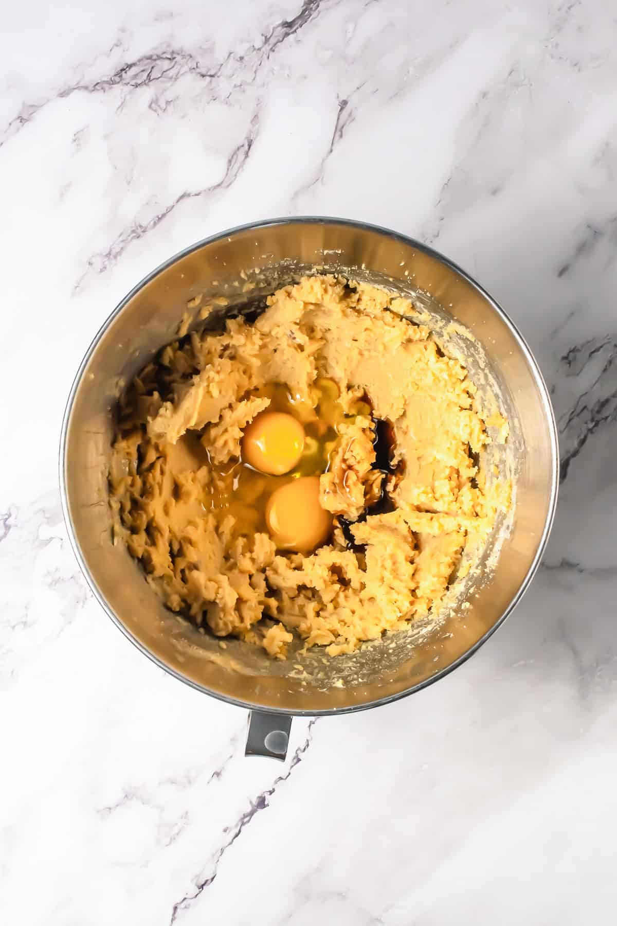 A mixing bowl with oatmeal cookie dough and two cracked eggs sits on a marble countertop, ready to be turned into delicious chocolate chip bars.