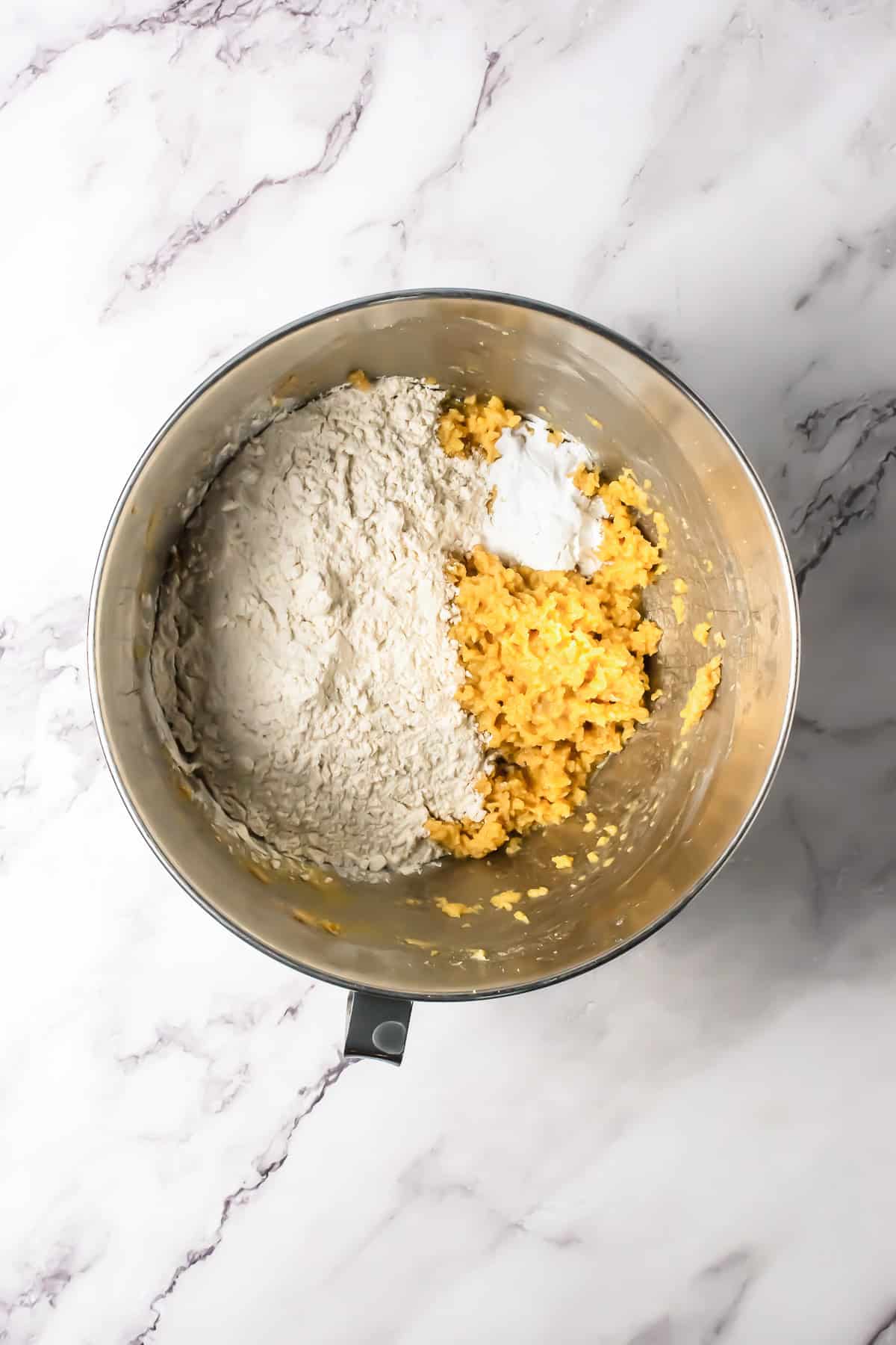 A metal mixing bowl with flour, baking powder, mashed bananas, and chocolate chips sits on a marble countertop.
