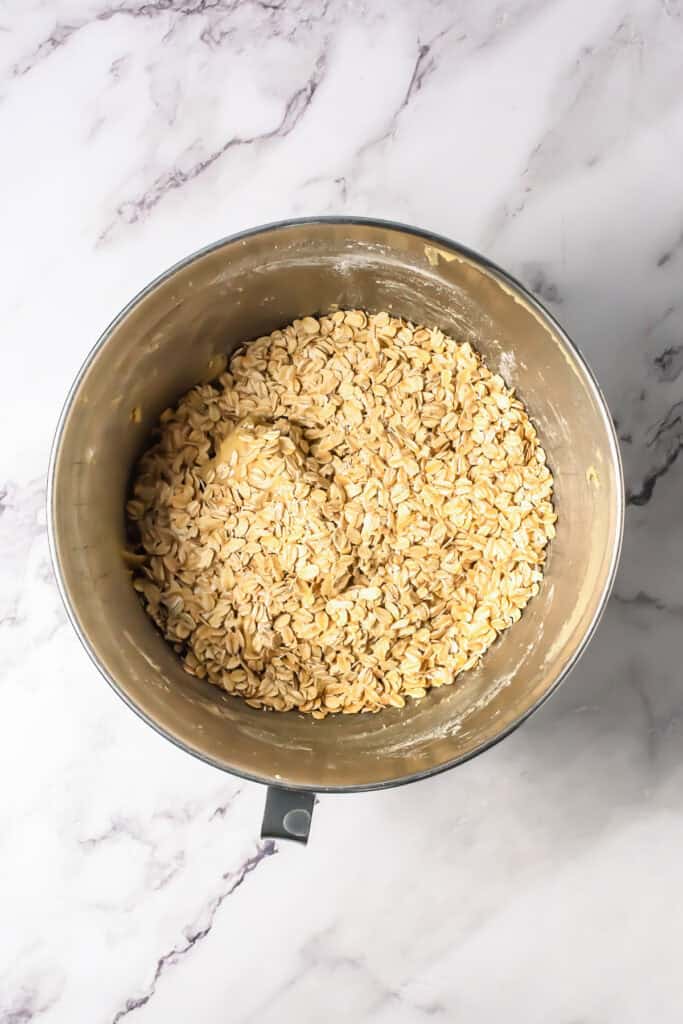 A metal mixing bowl filled with chocolate chip oatmeal cookie dough rests on a white marble surface.