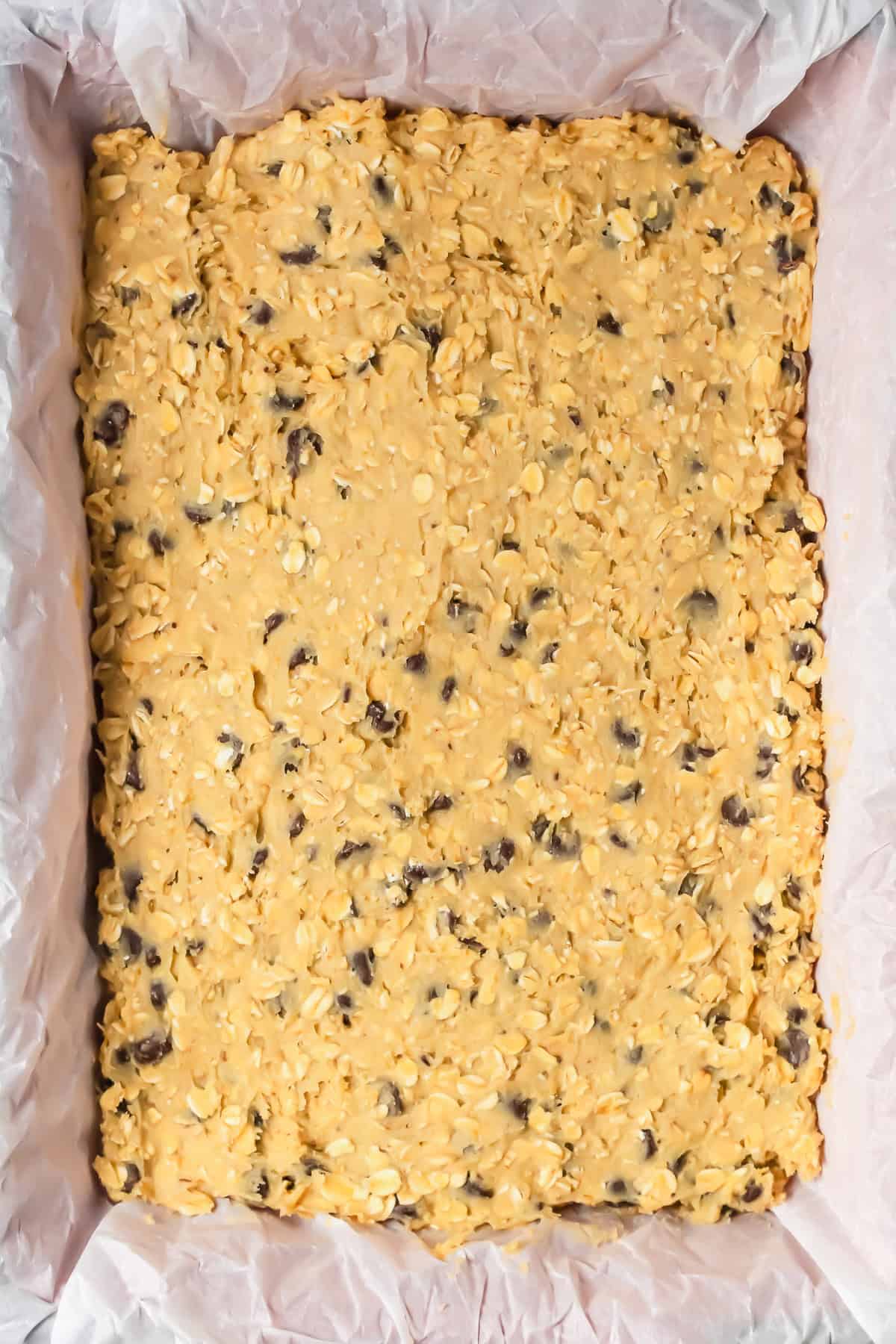 Unbaked oatmeal chocolate chip bars batter spread evenly in a parchment-lined rectangular baking dish.