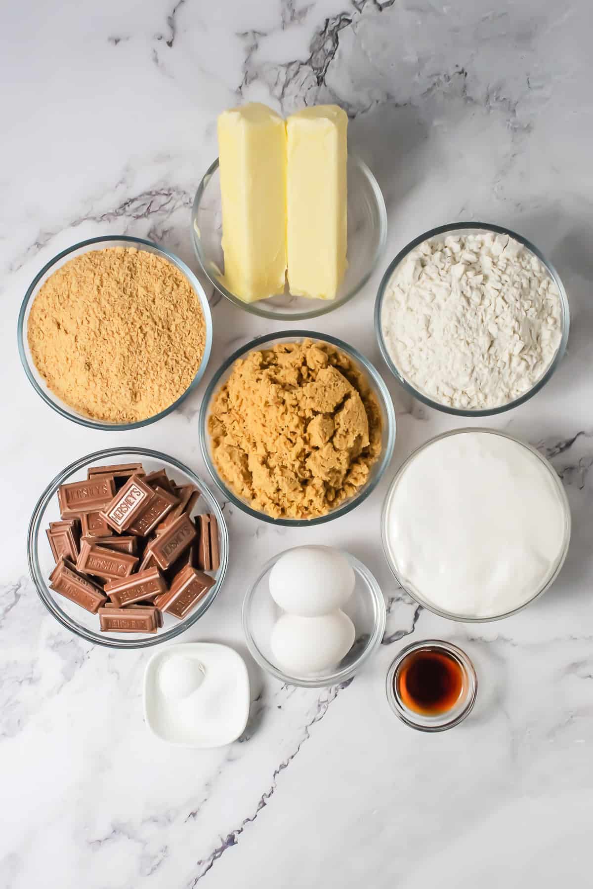 Baking ingredients for a delicious S&rsquo;mores Bars Recipe are arranged on a marble surface: butter, flour, sugar, eggs, chocolate, vanilla, and baking soda in bowls.