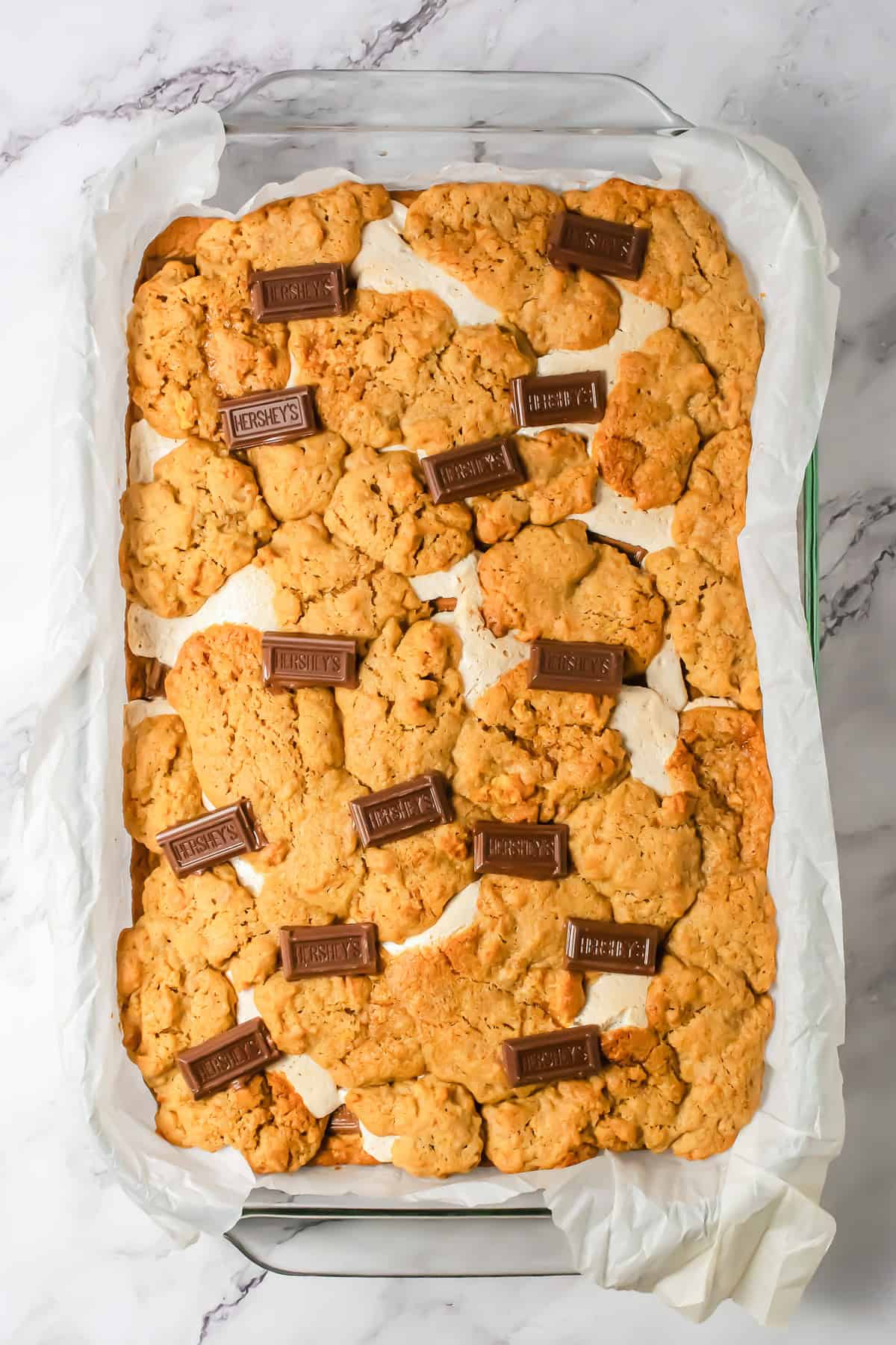 A baking dish of S&rsquo;mores Bars from a delicious S&rsquo;mores Bars Recipe, topped with oat cookie crumble and Hershey&rsquo;s chocolate pieces on parchment paper&mdash;an irresistible dessert recipe for any occasion.