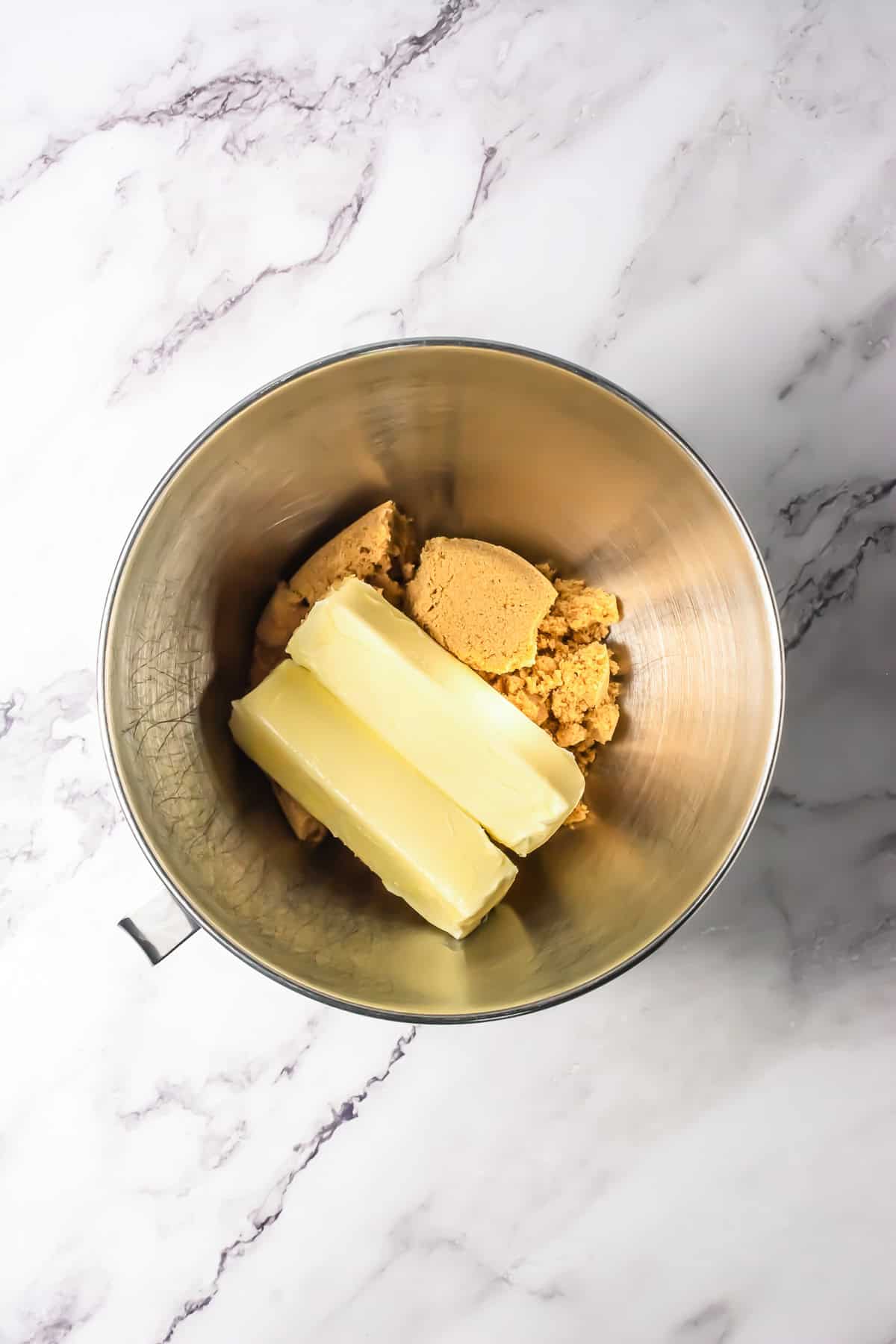 Two sticks of butter and brown sugar chunks in a metal mixing bowl on a marble surface, ready to be mixed for the ultimate S&rsquo;mores Bars Recipe.