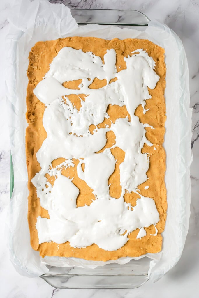 A baking dish with cookie dough pressed flat and uneven marshmallow spread on top, lined with parchment paper&mdash;perfect for making a delicious S&rsquo;mores Bars Recipe.