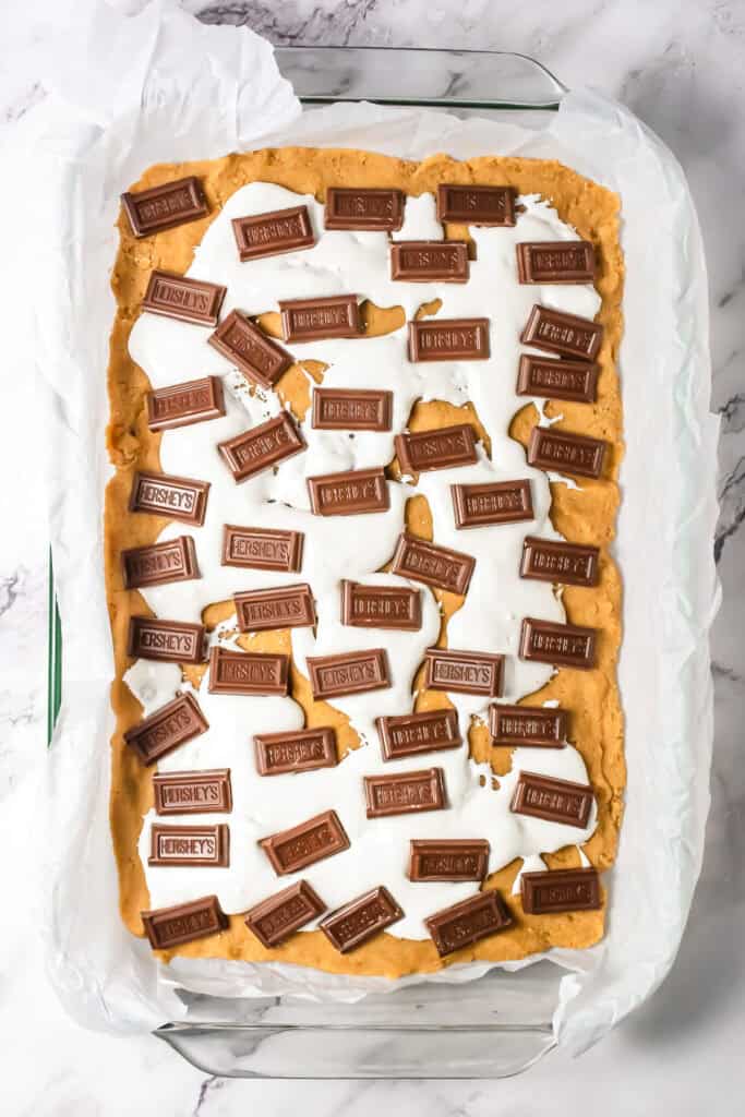 A baking dish with cookie dough, melted marshmallow, and Hershey's chocolate pieces on top, this S&rsquo;mores Bars dessert is ready to bake.