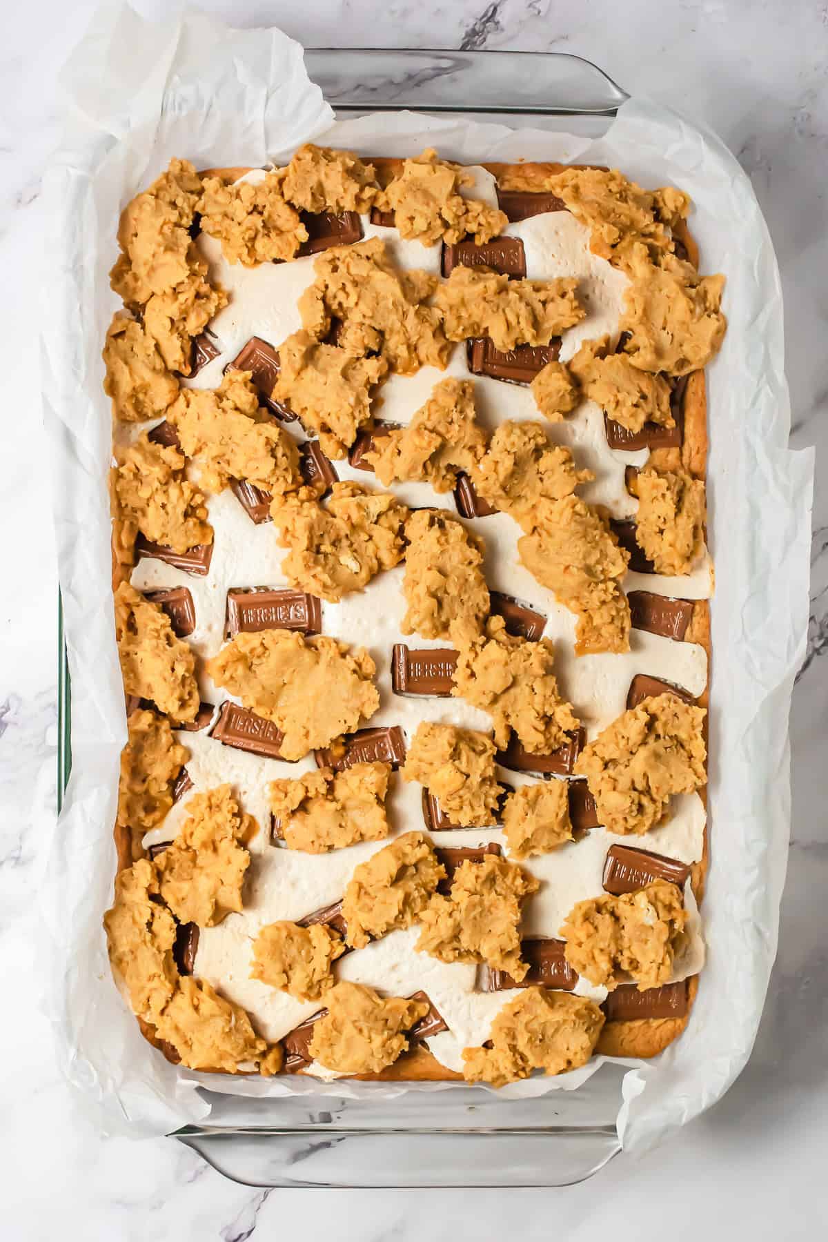 Rectangular pan lined with parchment paper, topped with marshmallows, chocolate pieces, and dollops of cookie dough&mdash;perfect for a classic S&rsquo;mores Bars Recipe everyone will love.