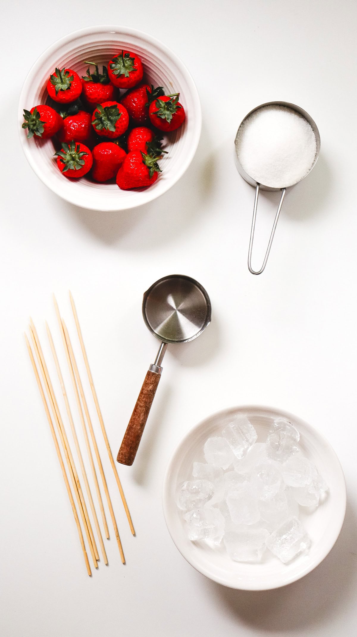 Top view of ingredients for a delicious Strawberry Tanghulu: fresh strawberries, a cup of sugar, ice cubes, skewers, and a metal scoop, all arranged on a white background.