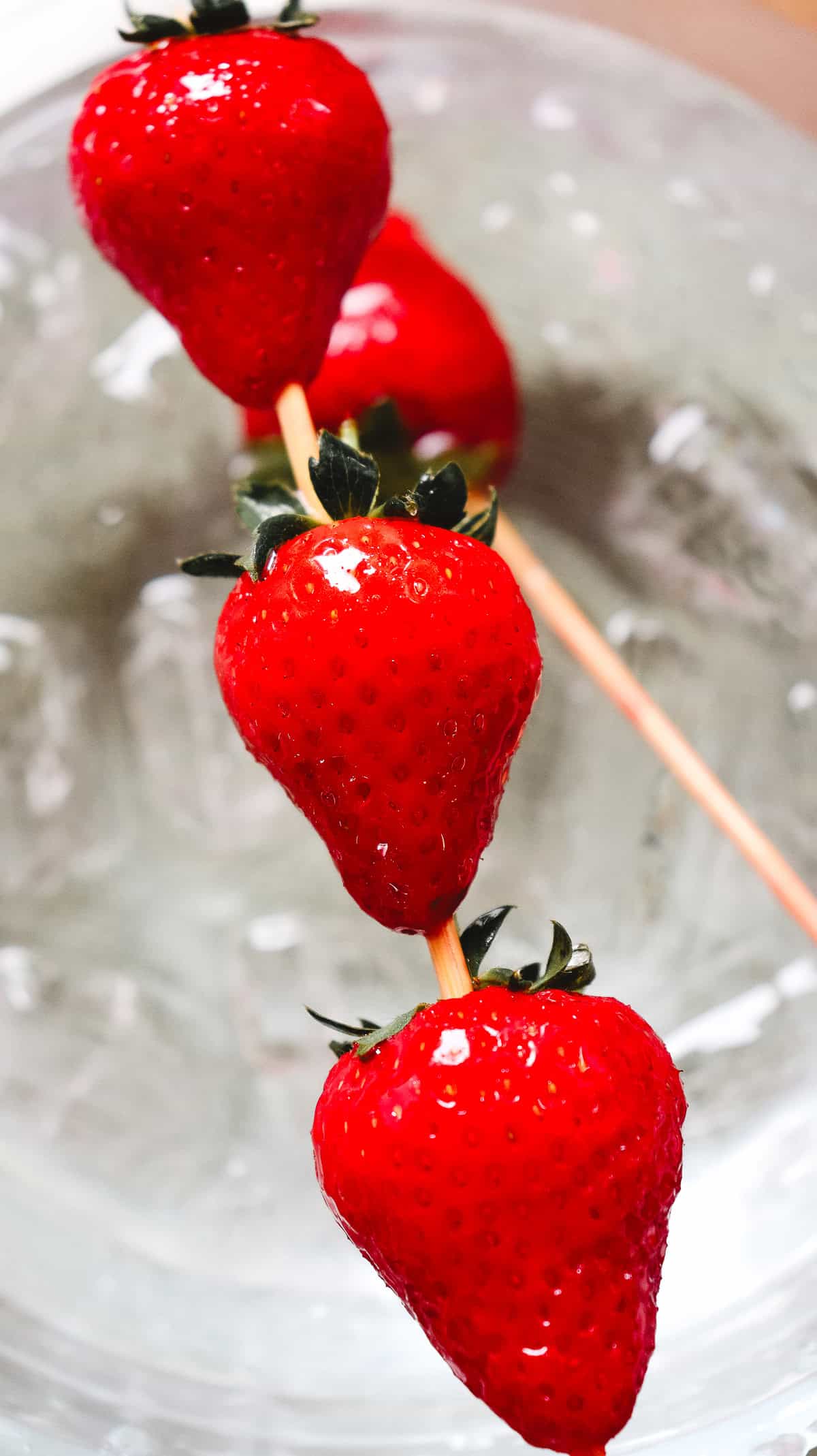 Four fresh strawberries on wooden skewers rest over a glass bowl filled with ice&mdash;perfect for making a classic Strawberry Tanghulu or trying out your favorite Tanghulu Recipe at home.