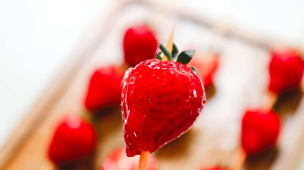 A close-up of a glossy strawberry tanghulu on a skewer, with more strawberries blurred in the background&mdash;perfect inspiration for your next strawberry tanghulu recipe.