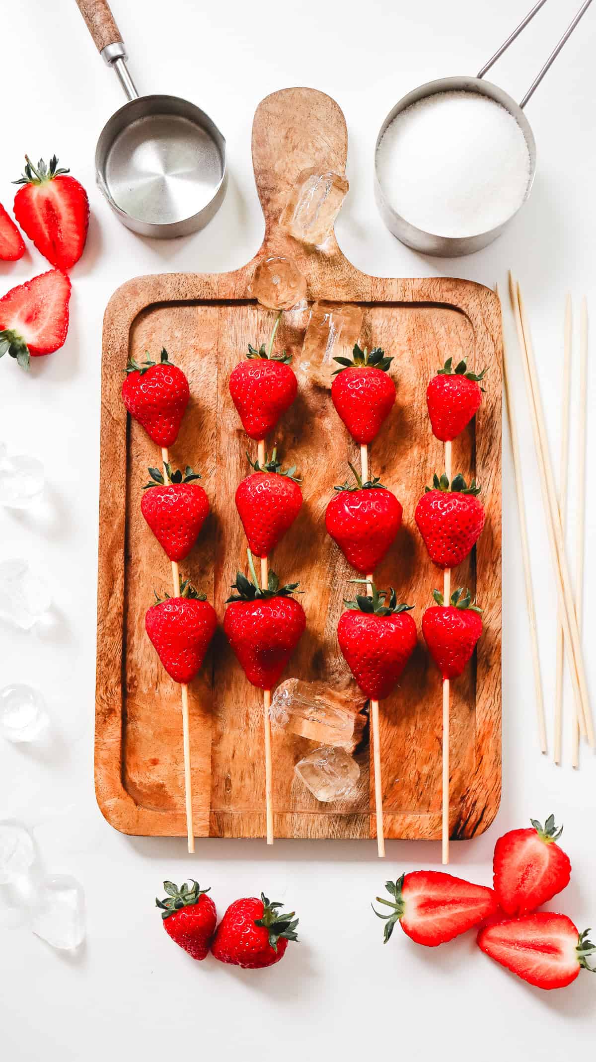 Four skewers with whole strawberries on a wooden board, surrounded by sugar, cut strawberries, and ice cubes&mdash;perfect for trying your own Strawberry Tanghulu recipe at home.