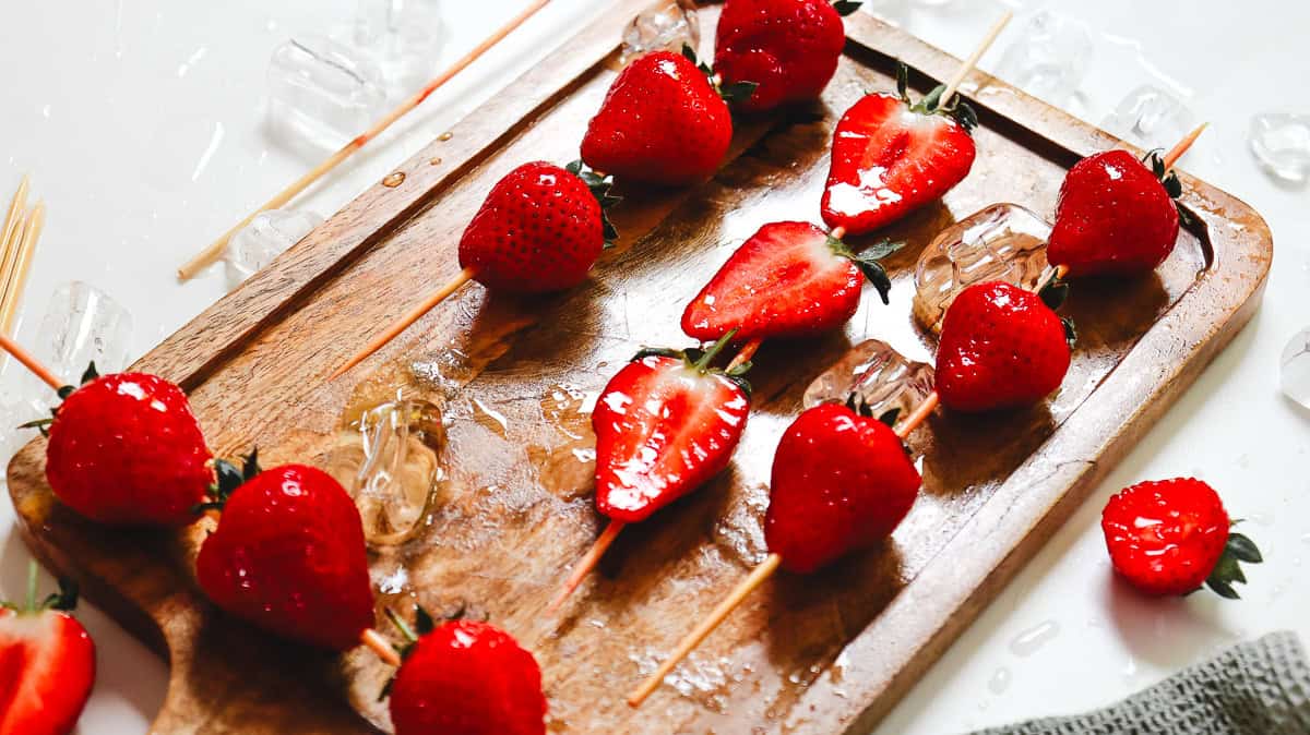 Strawberries and ice cubes on wooden skewers create a refreshing Strawberry Tanghulu, displayed on a wooden cutting board with scattered ice around.