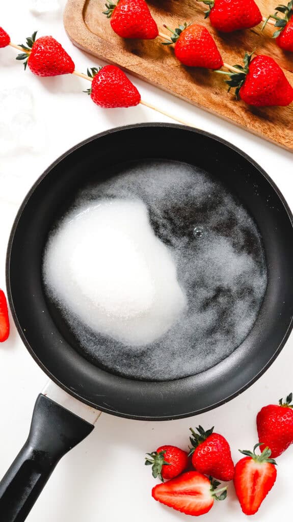 A pan with sugar dissolving in water, surrounded by fresh strawberries on a white surface, perfect for making a classic Strawberry Tanghulu recipe.