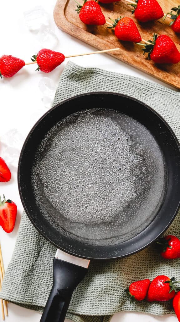 A frying pan with bubbling water sits on a towel, surrounded by skewered and loose strawberries&mdash;perfect for crafting homemade Strawberry Tanghulu.