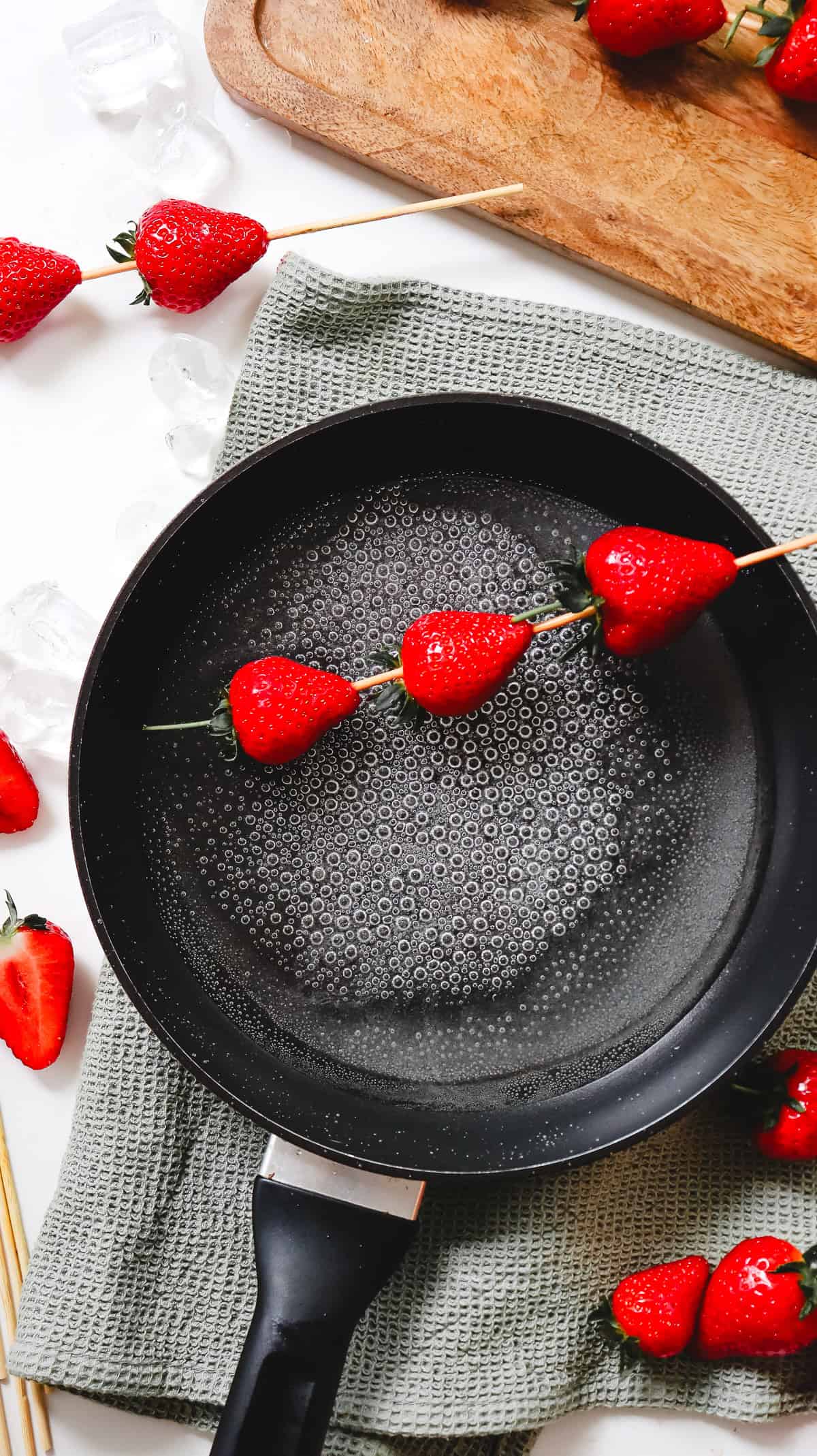 Fresh strawberries on a skewer held over a nonstick pan filled with hot water, surrounded by ice and more strawberries&mdash;perfect for creating a classic Strawberry Tanghulu recipe.