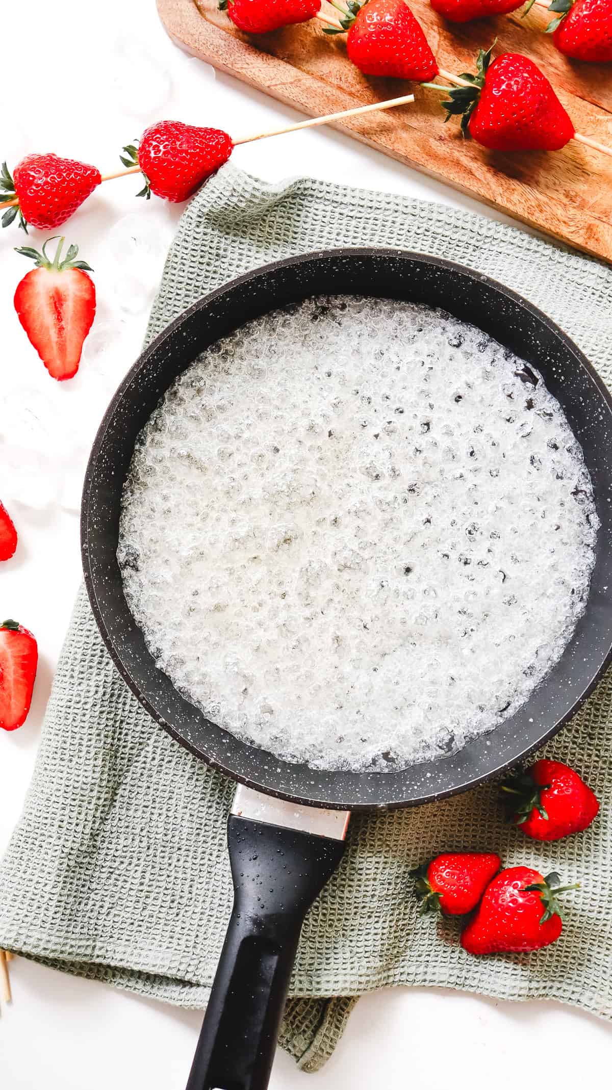 A saucepan with bubbling sugar syrup sits on a towel, surrounded by fresh strawberries and a cutting board&mdash;a perfect setup for making Strawberry Tanghulu or trying your favorite Tanghulu Recipe.