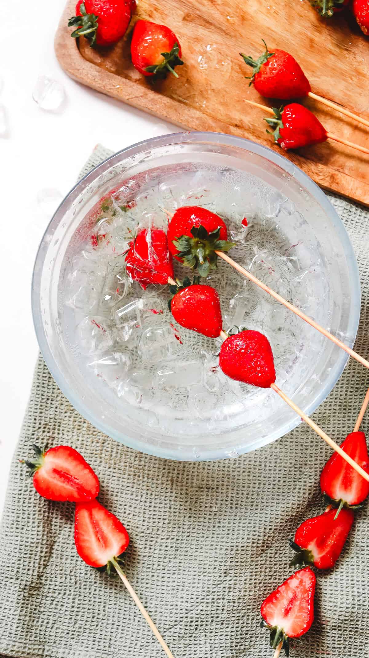 Skewered strawberries in a bowl of ice water, perfect for a Strawberry Tanghulu treat, with more strawberries on a cutting board and cloth nearby.