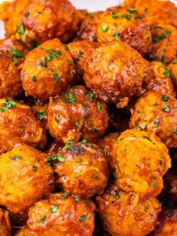 A white plate piled high with saucy, garnished meatballs, with a fork and shredded cheese nearby.