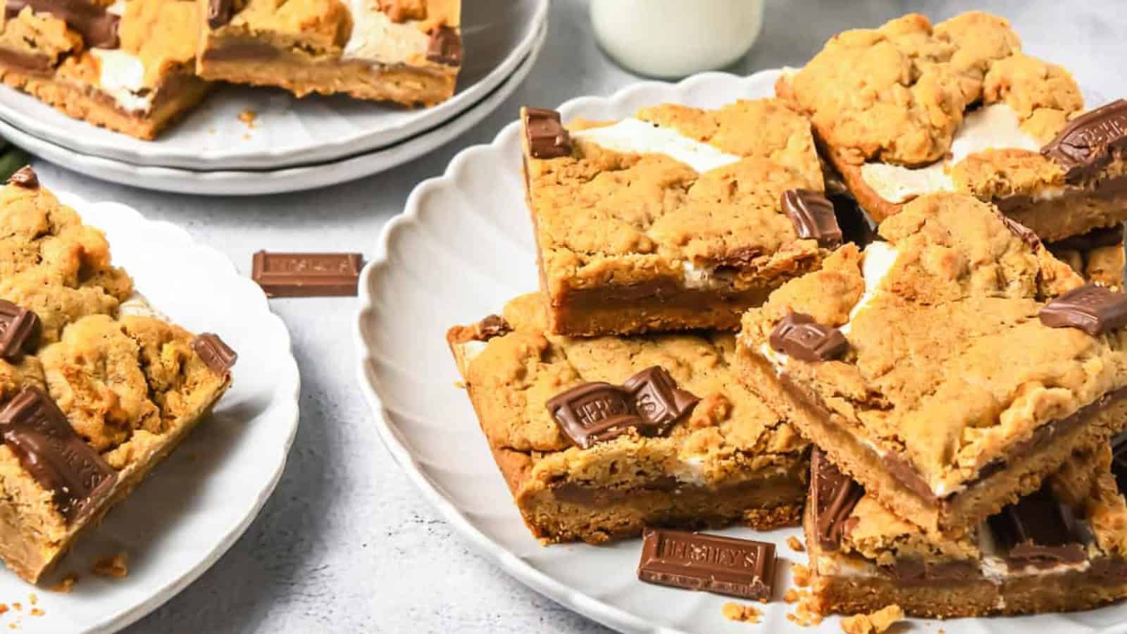 Plates of s’mores cookie bars with chocolate pieces and a glass of milk in the background.