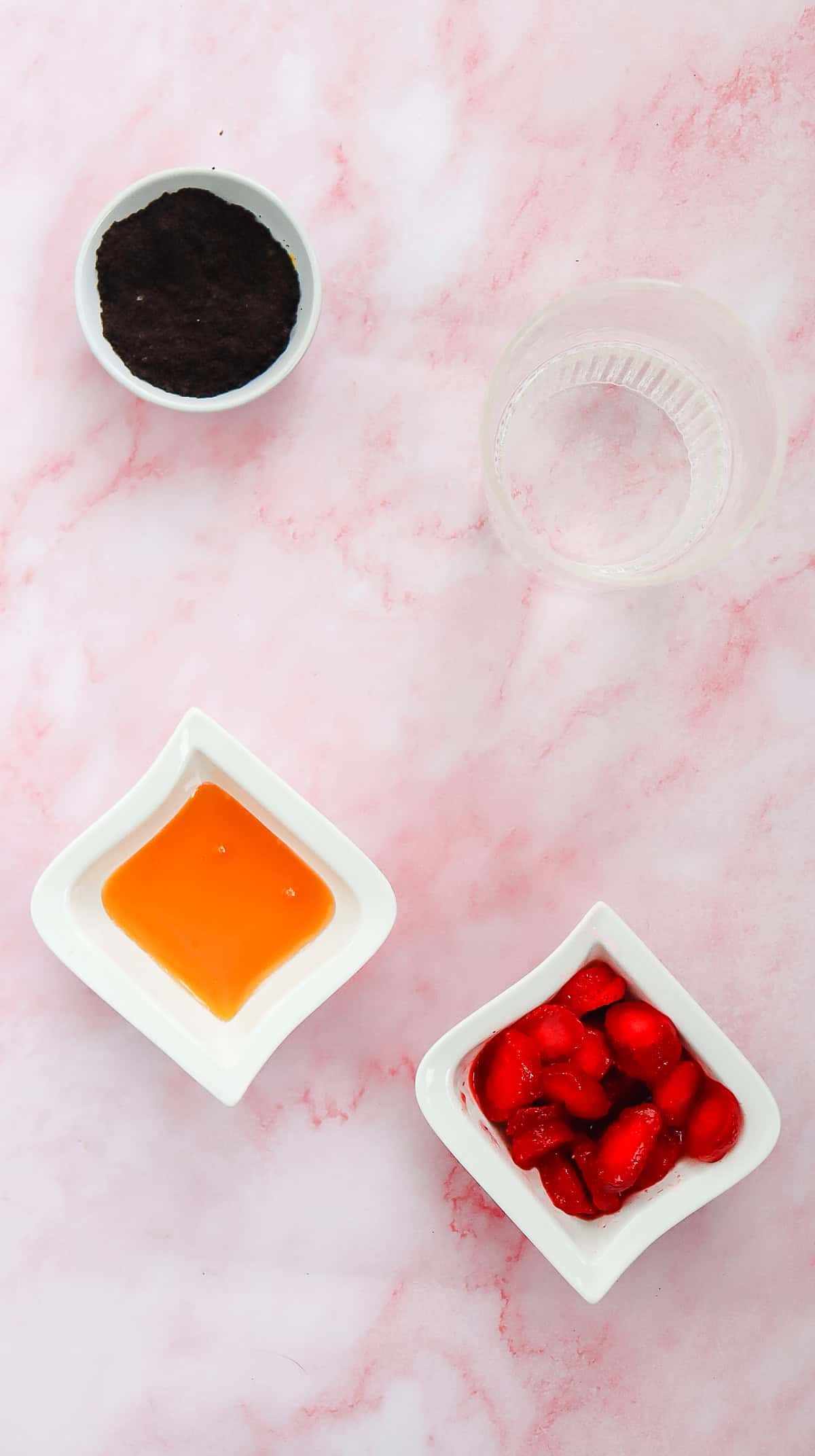 Four small bowls with cocoa powder, clear liquid, orange syrup, and sliced red fruit on a pink marble surface&mdash;perfect inspiration for your own Starbucks Refresher or Strawberry Acai Refresher copycat at home.