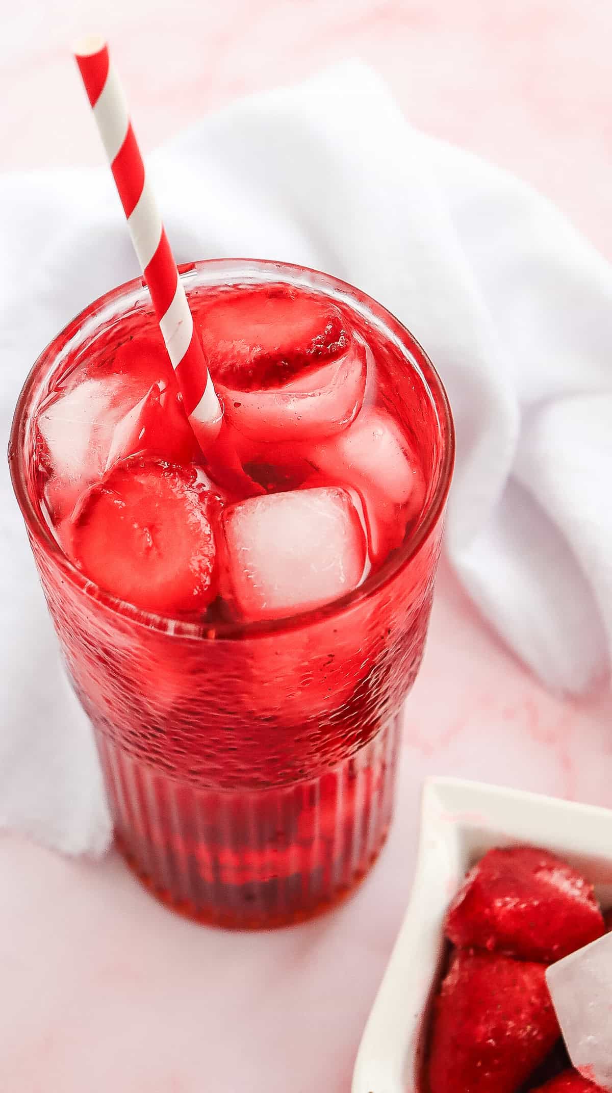 A glass of red iced drink, inspired by a Starbucks Copycat Strawberry Acai Refresher, with a red and white striped straw on a pink surface, next to a white cloth.