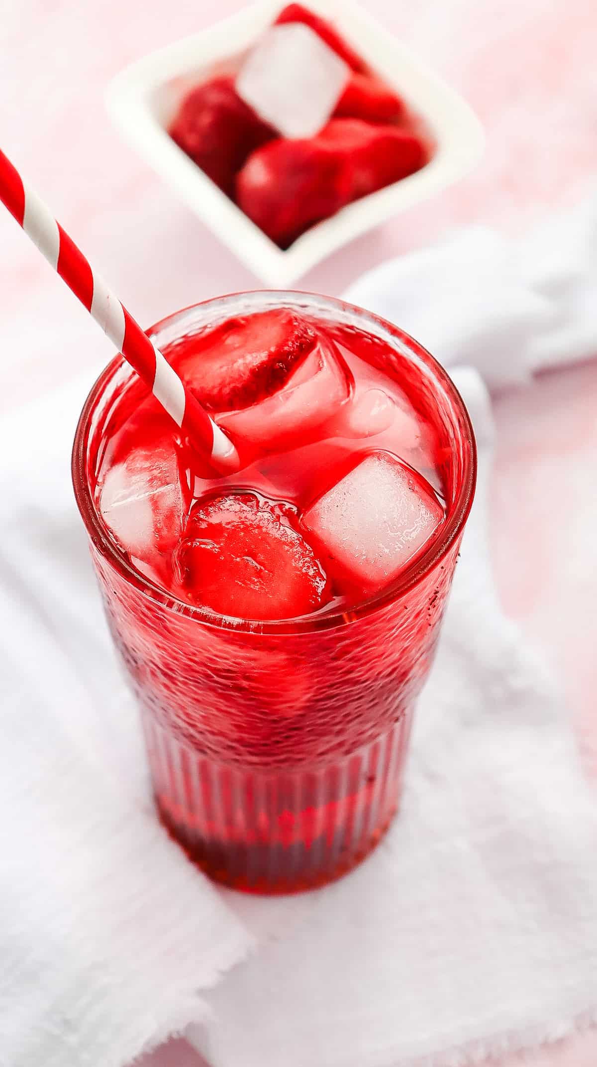 A glass of red Strawberry Acai Refresher with ice and a striped straw, with red ice cubes in a bowl in the background&mdash;perfect for a Starbucks copycat experience at home.
