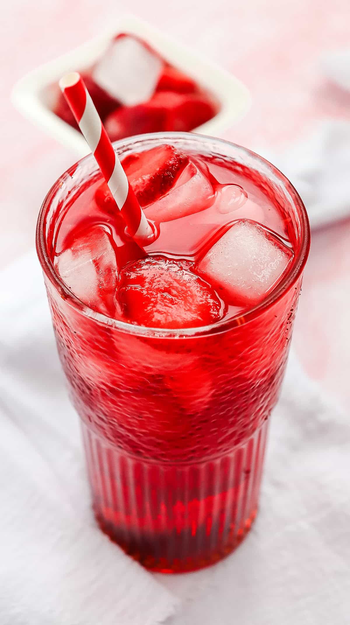 Red drink with ice cubes in a glass, striped straw, napkin underneath, and ice tray in the background&mdash;this vibrant Strawberry Acai Refresher is a perfect Starbucks Copycat for refreshing summer days.