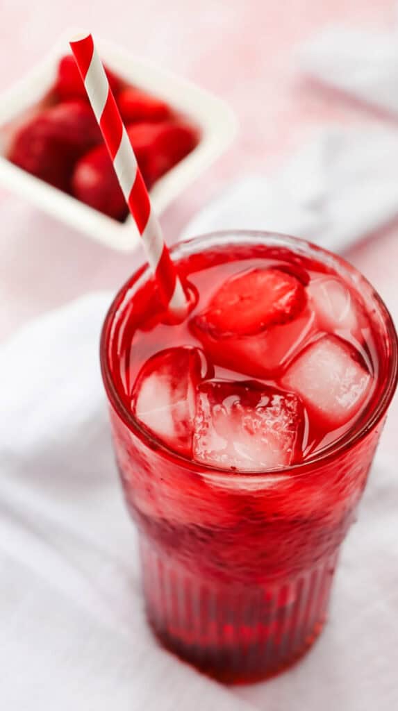 Glass of red strawberry drink with ice cubes and a red-striped straw, inspired by the Strawberry Acai Refresher, with fresh strawberries in the background for a true Starbucks Copycat vibe.