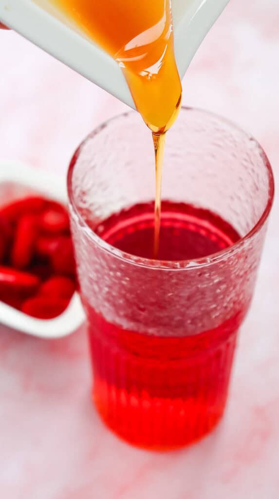 Golden liquid being poured into a textured glass filled with a vibrant red Strawberry Acai Refresher, Starbucks copycat style, with red berries in the background.