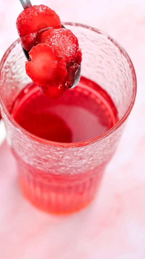 A spoon with sliced strawberries held above a glass of vibrant red Starbucks Copycat Strawberry Acai Refresher on a pink background.