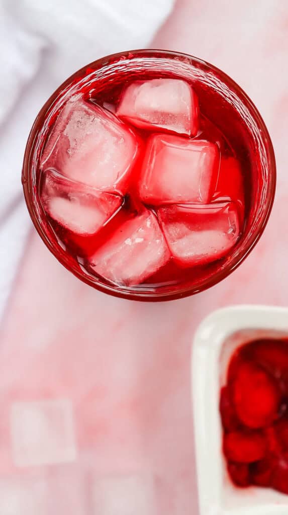 A glass of vibrant Strawberry Acai Refresher filled with ice cubes seen from above on a light pink surface&mdash;perfect for a Starbucks Refresher copycat experience at home.