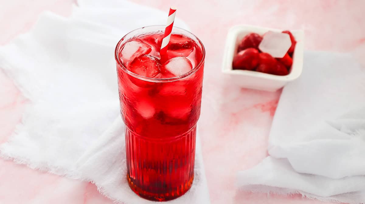A tall glass of red iced drink with a striped straw, next to a bowl of cherries on a white cloth&mdash;a tempting copycat Starbucks Strawberry Acai Refresher perfect for summer sipping.
