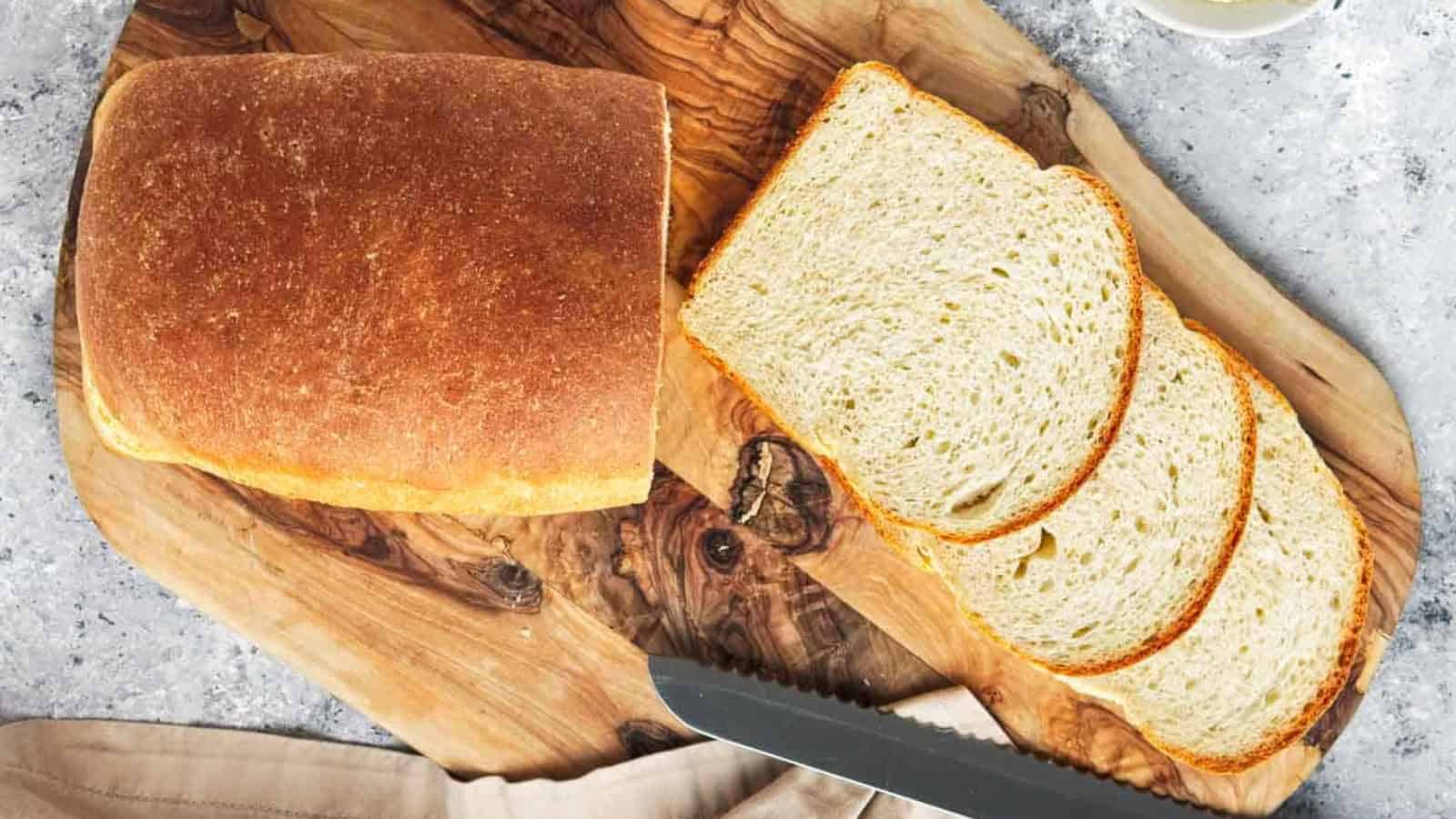 A loaf of bread with three slices cut, placed on a wooden cutting board with a bread knife nearby.