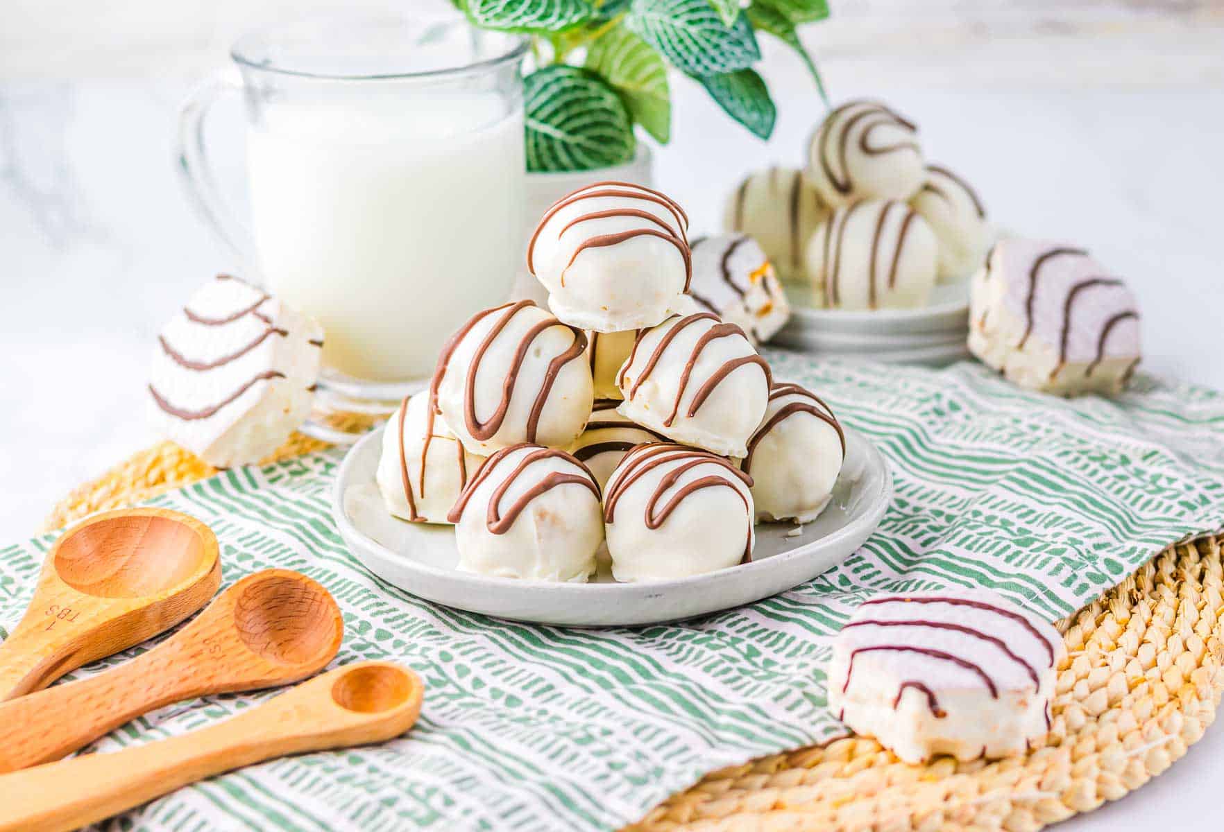Zebra Cake Balls: White chocolate-covered cookies with chocolate drizzle on a plate, paired with a glass of milk and measuring spoons nearby.