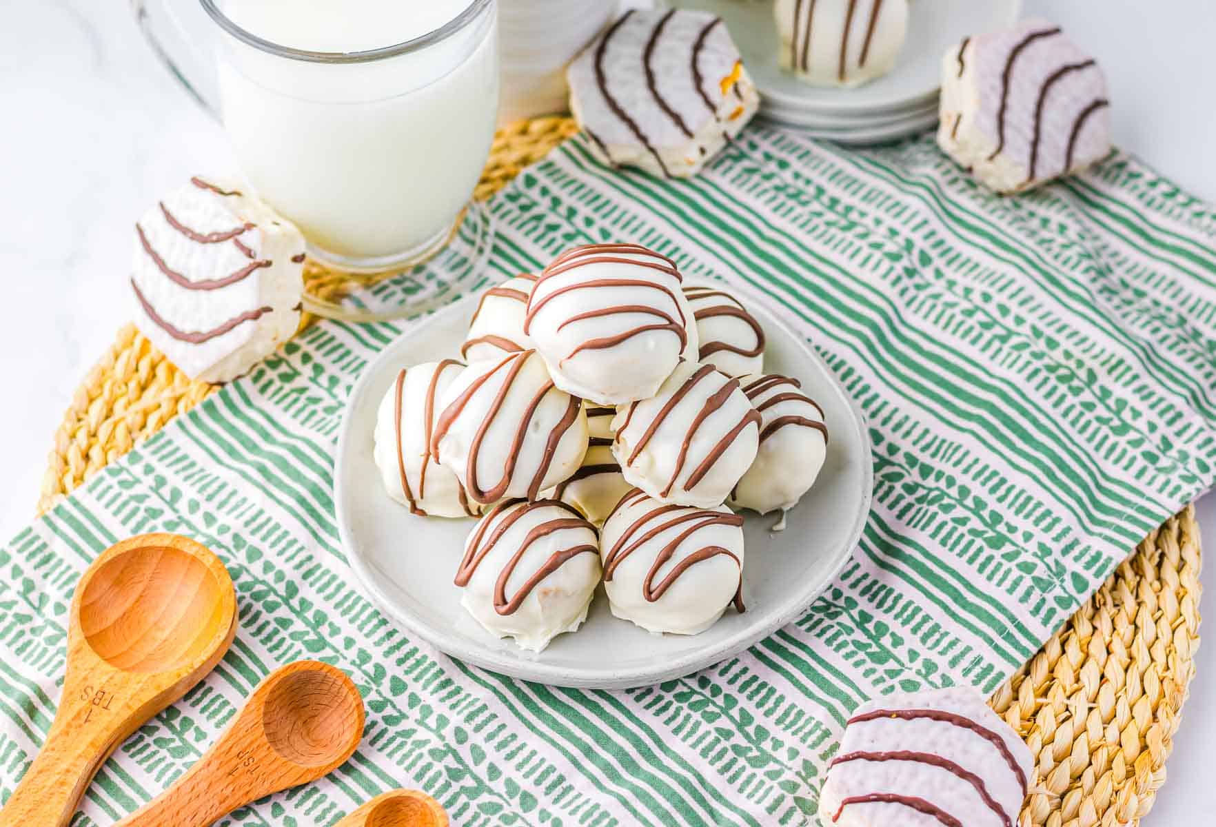 A plate of white chocolate cookies with chocolate drizzle, reminiscent of Zebra Cake Balls, sits next to a glass of milk and wooden measuring spoons.