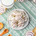 Plate of Zebra Cake Balls covered in white chocolate with a chocolate drizzle, next to a glass of milk and wooden spoons on a placemat.