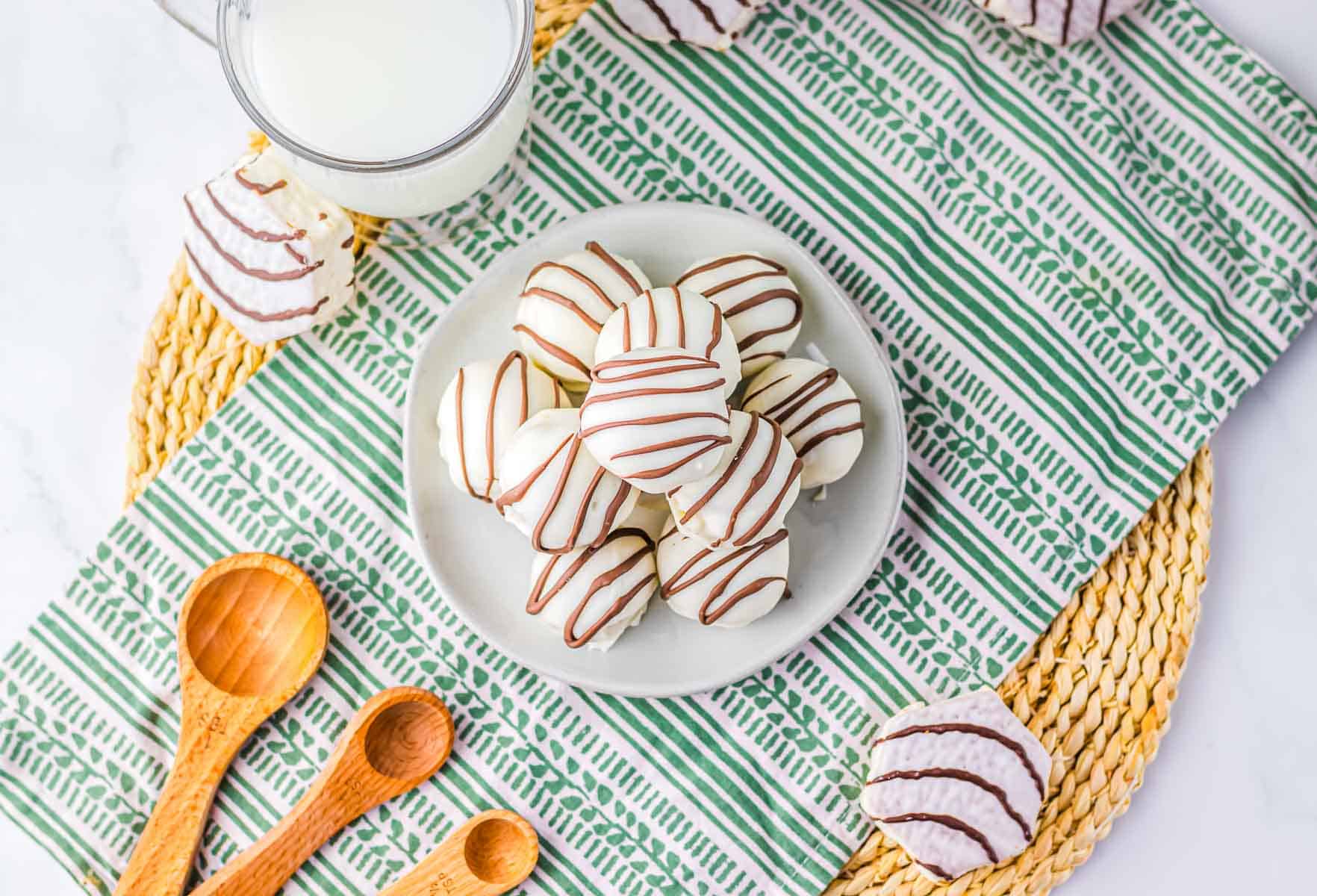 White chocolate truffles with chocolate drizzle, resembling Zebra Cake Balls, arranged on a plate next to a glass of milk and wooden measuring spoons.