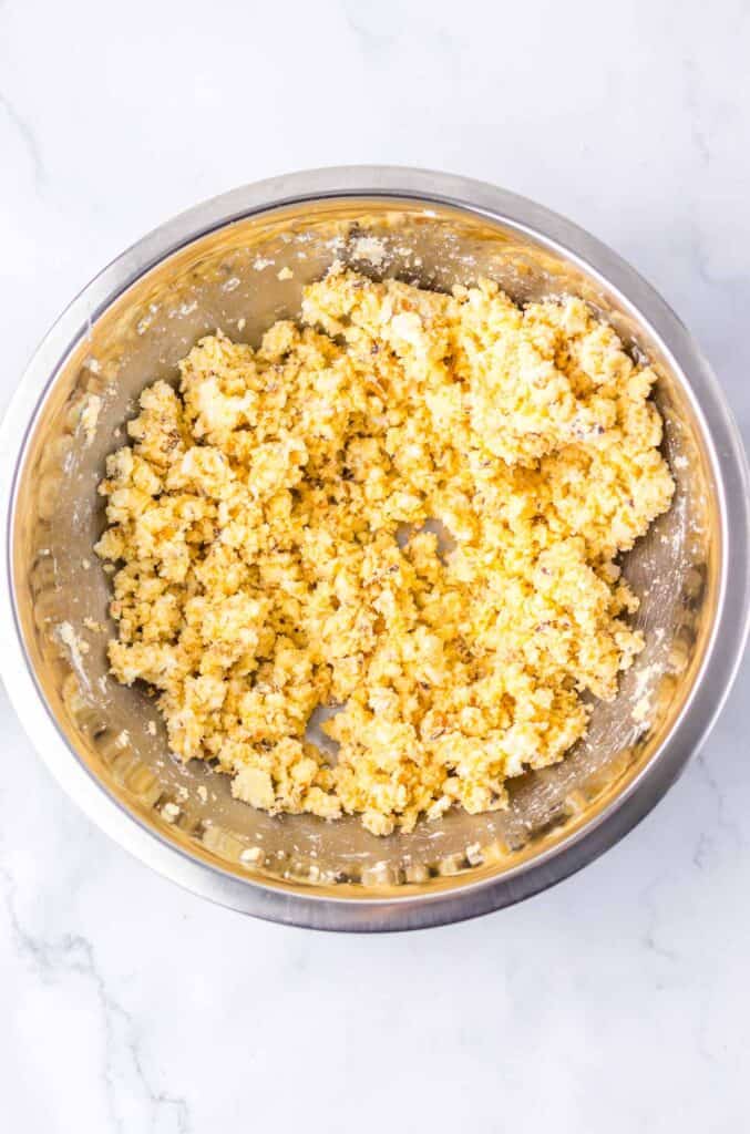 A stainless steel bowl filled with crumbly yellow biscuit dough mixture for Zebra Cake Balls sits on a white marble surface.