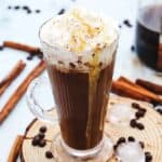 Glass mug of Copycat Starbucks Iced Cinnamon Dolce Latte topped with whipped cream and caramel, surrounded by cinnamon sticks and coffee beans.